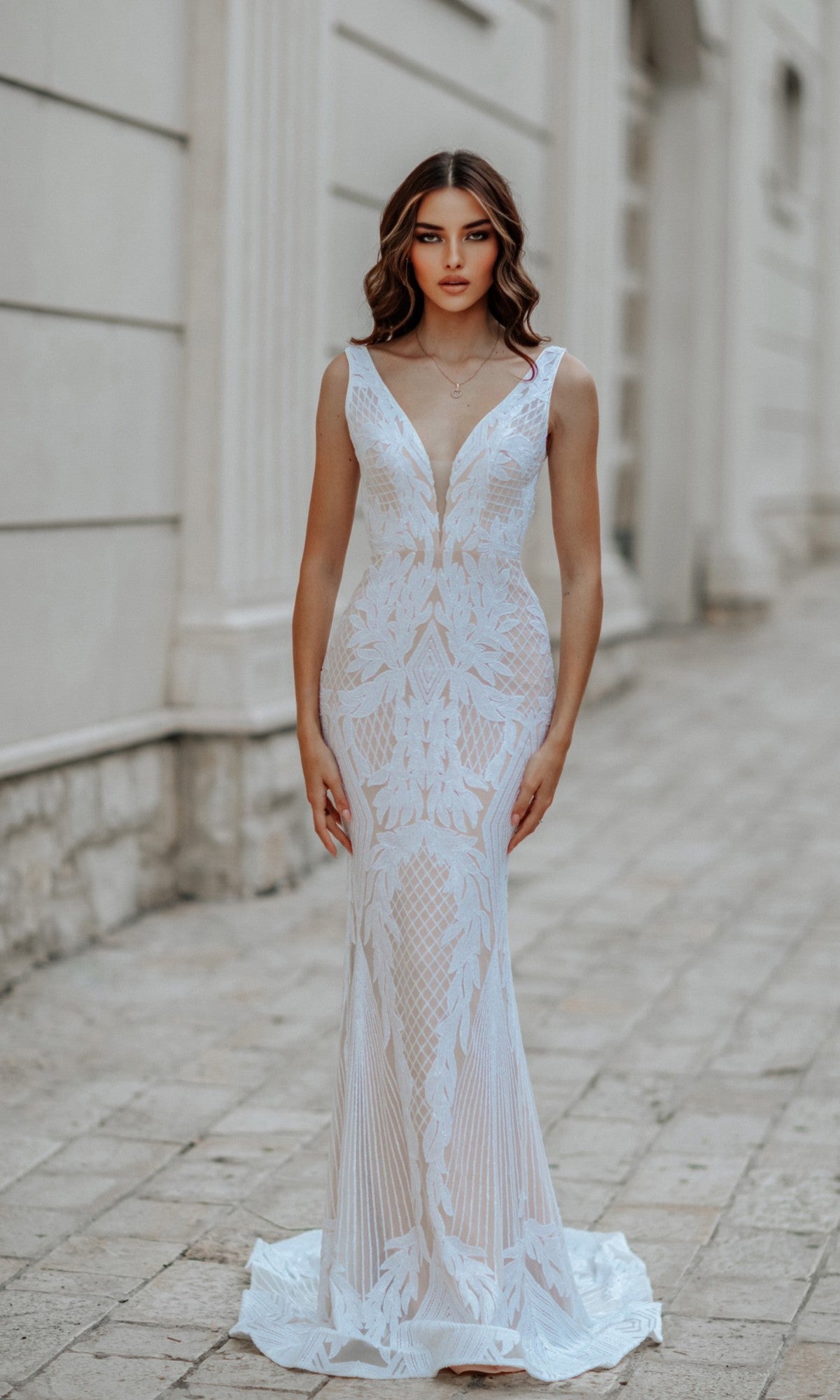 A woman stands outside wearing the Classic Long White Formal Dress: Tina Holly BB052, a sleeveless, form-fitting mermaid gown with white lace, a deep V neckline, and geometric patterns.